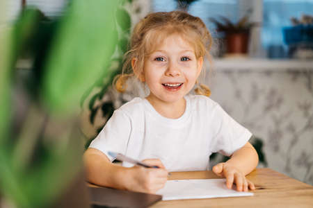 little cute blonde girl writes, studies at home in front of a laptop, online learning, back to schoolの写真素材