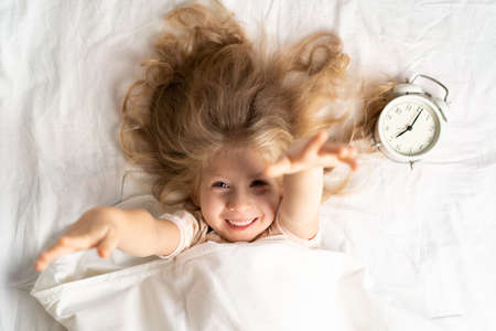 happy little blonde girl in a white bed with an alarm clock, good morningの写真素材