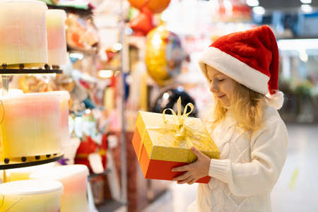 a little girl in a Christmas hat with a New Years gift in her hands at a showcase with toys. children choose gifts for Christmas. Black Friday discountsの写真素材
