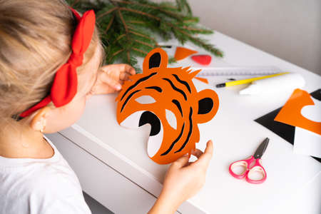 a little girl in a Christmas hat makes a New Years tiger mask from colored paper, the symbol of 2022, with her own handsの写真素材