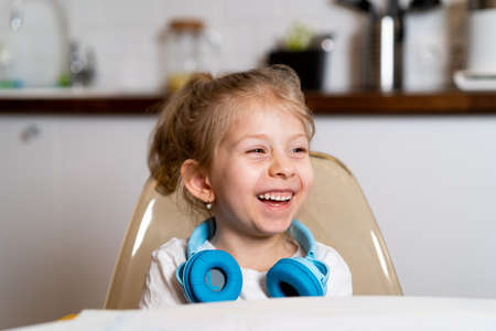 A little blonde girl at home in the kitchen at the table with headphones draws and learns remotely...