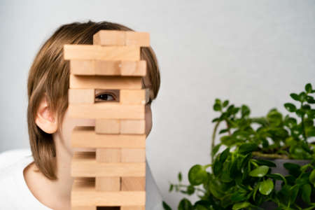 a happy boy is playing a board game, building a tower of wooden cubes, hiding behind a tower of...