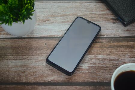 Blank screen smartphone between coffee cup an vase on wood tableの写真素材