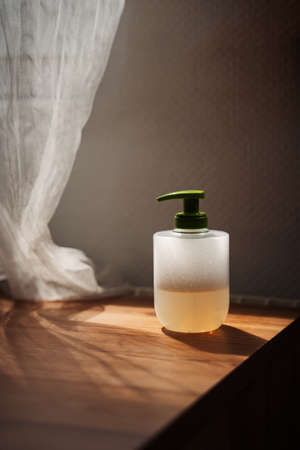 Liquid hand soap with foam and a green cap on wooden table with backlit curtainの写真素材