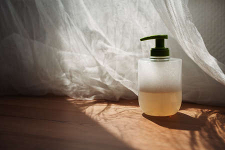 Liquid hand soap with foam and a green cap on wooden table with backlit curtainの写真素材