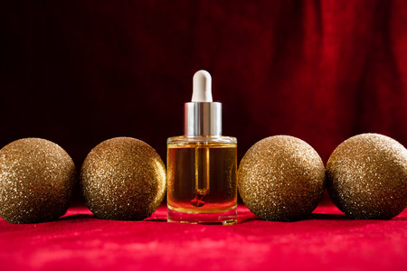 Gold glitter christmas balls in a row and face oil in glass bottle with dropper on red velvet with moody lighting background. Classic holiday colors, luxury festive atmosphere and self-careの写真素材