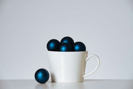 Dark blue Christmas balls baubles in white cup on white background. Modern minimalism holiday decoration concept. Front view.の写真素材