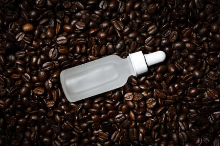 Face serum in matte white glass bottle with dropper on whole roasted coffee bean textured...