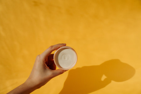 Female Hand holding white cream in a beige pot on sunlit yellow background with textured shadows. Moisturising sunscreen product for summer holidaysの写真素材