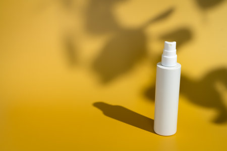 Blank white spray bottle on yellow background in direct sunlight with plant leaf shadow. Summer skin...
