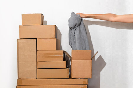 Pile of Cardboard Boxes in Minimalist Setting and female hand holding clothes, representing concepts...