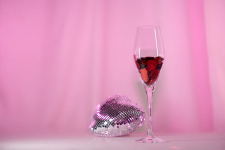 Wine glass with heart confetti and disco on pink background. Minimalist holiday photo for Valentinesの写真素材