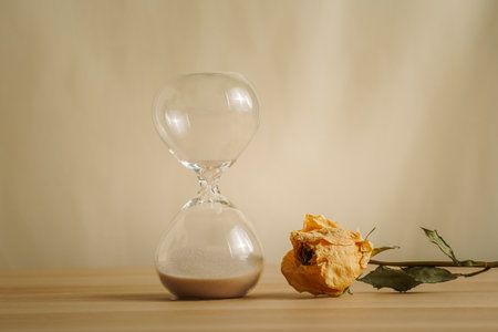 A simple hourglass on a wooden table with dry rose a beige backdrop, symbolizing time flow, aging,...