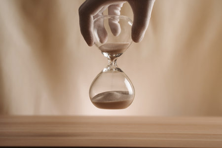 A glove-covered hand holding an hourglass on a wooden surface, symbolizing the flow of time,...