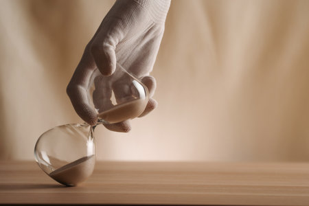 A glove-covered hand holding a tipped hourglass on a wooden surface, symbolizing the flow of time,...