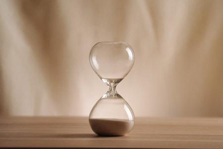 A simple hourglass on a wooden table with a beige backdrop, symbolizing time flow and minimalistic...