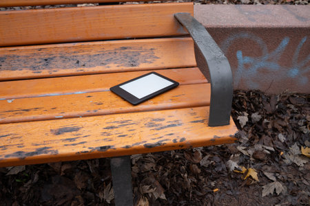 Ebook resting on a light wooden park bench outdoors. Everyday scene showing a personal object, representing reading, leisure, and casual outdoor life.の写真素材