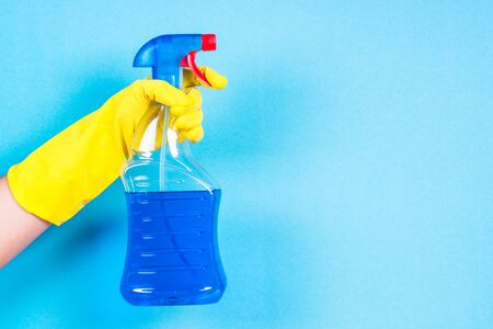 Hand in yellow gloves hold cleaning agent bottle on blue background.