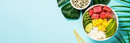 Tuna poke bowl with rice, avocado, mango and cucumber on blue.