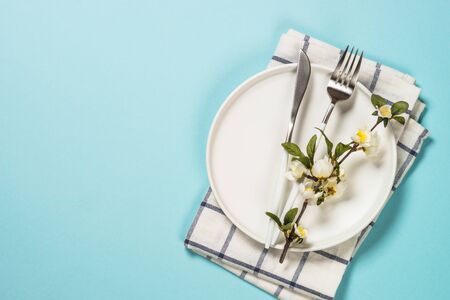 Spring table setting with blossom flower, white plate and cutlery on blue background. Flat lay...