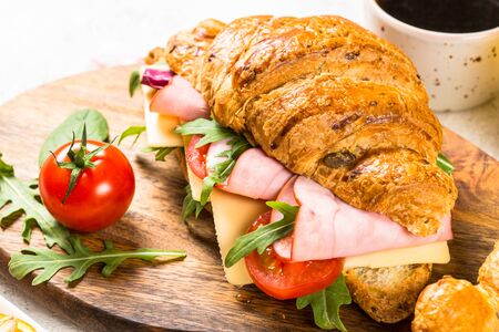 Croissant sandwich with ham, cheese and vegetables on wooden cutting board with cup of coffeee. Healthy snack or breakfast.の写真素材