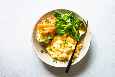 Scrambled eggs, sandwich with cream cheese and salmon and salad leaves at white stone table.の写真素材
