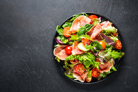 Green salad on black background. Green salad leaves, smoked meat and tomatoes. Top view with space for text.の写真素材