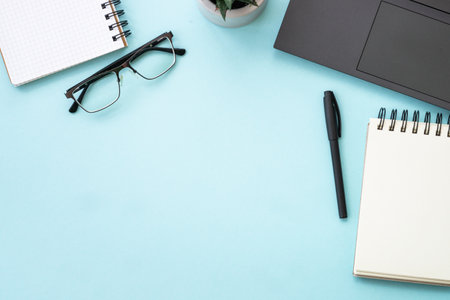 Office desk with laptop, notepad, pen and glasses. Flat lay image on blue with copy space.の写真素材