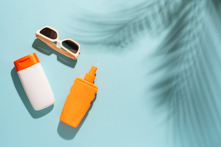 Sunscreen and sunglasses on blue background with hard sunny shadows. Flat lay with copy space.