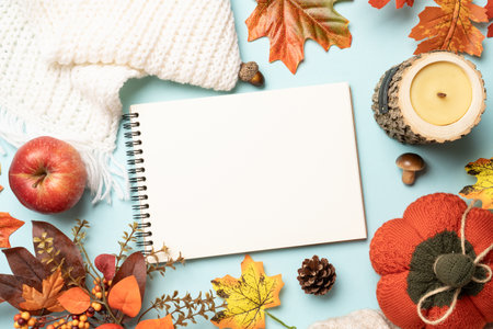 Autumn flat lay background, falling leaves, notepad, knitted scarf and wooden candle on blue...