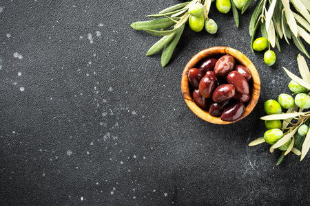 Olives in wooden bowl and fresh olive tree branch at black background. Top view with space for design.の写真素材