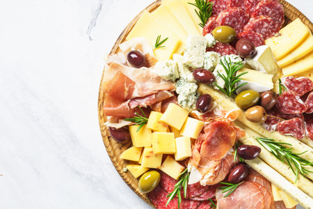 Antipasto platter. Cheese and meat - jamon, salami, olives at wooden board at marble table. Top...