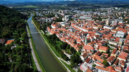 Scenic aerial panorama of a charming European city with red rooftops, a flowing river, lush greenery, and distant mountain ranges under a clear blue skyの写真素材