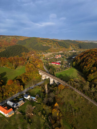 A picturesque mountain valley in autumn with colorful forests, a historic railway bridge, small houses, and rolling hills under a calm skyの写真素材