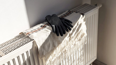 Black gloves and a wool scarf placed on a home radiator, symbolizing heating, winter warmth,...