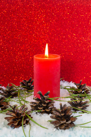 Burning candle in the snow surrounded by cones and fir needles on a red background, new year mood, front view, copy spaceの写真素材
