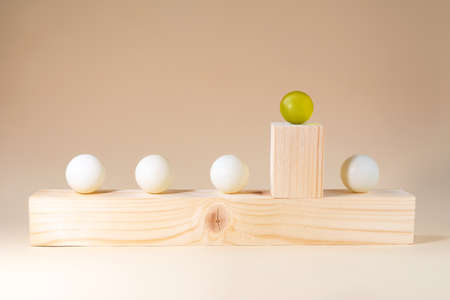 Outstanding yellow sphere among white spheres on a wooden bar on a pastel-beige background. Difference in color and height. Minimal conceptual ideaの写真素材