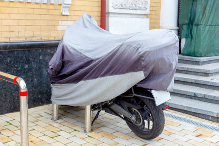 The motorcycle is on the street, covered with a gray cover, near the house and stairsの写真素材