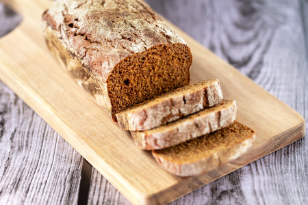 Loaf of freshly baked dark bread with sliced pieces on a board on a wooden background