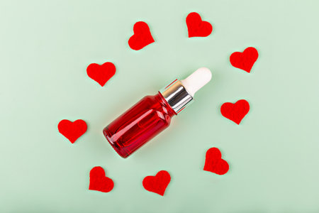 Red cosmetic bottle with dropper in circle of small red hearts on light green background. The concept of care, love, body careの写真素材