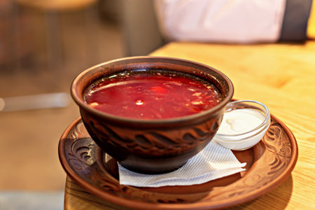 Hearty portion of classic borscht in carved clay bowl with sour cream in glass bowl, conveys homely...