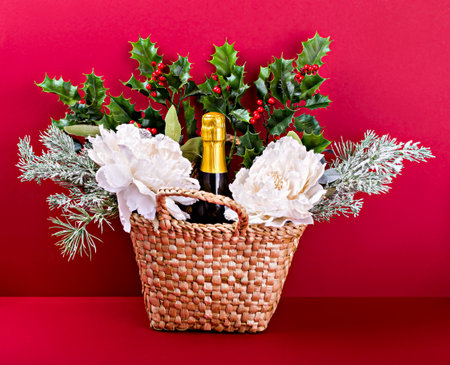 Wicker gift basket with bottle of sparkling wine, white flowers, holly and pine branches on red background. Ideal for holiday giftingの写真素材