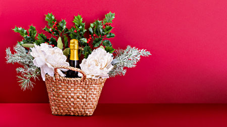 Wicker gift basket with bottle of sparkling wine, white flowers, holly and pine branches on red...