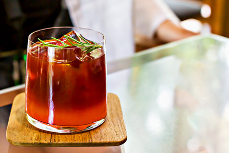 Glass of iced tea carcade or cocktail garnished with rosemary on wooden coaster, placed on reflective glass table with blurred background, creating relaxing atmosphereの写真素材