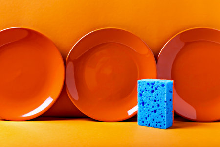 Three vibrant orange plates with contrasting blue sponge against an orange background, creating bold color composition. Perfect for cleaning and kitchen hygiene themesの写真素材