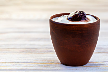 Clay cup filled with creamy sour cream topped or yogurt with spoonful of strawberry jam, placed on rustic wooden table. Simple and traditional dairy dessert yogurt or sour cream with homemade touchの写真素材