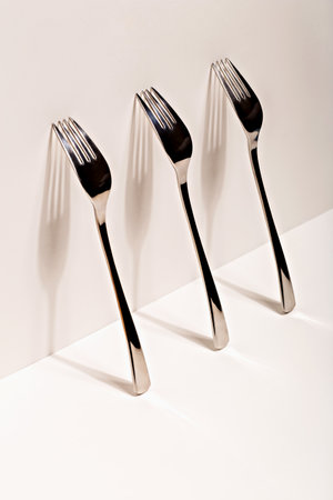 Three metallic forks leaning against white wall, casting dramatic elongated shadows. Composition highlights symmetry, contrast and light play. Concept of design, reflection and abstract aestheticsの写真素材