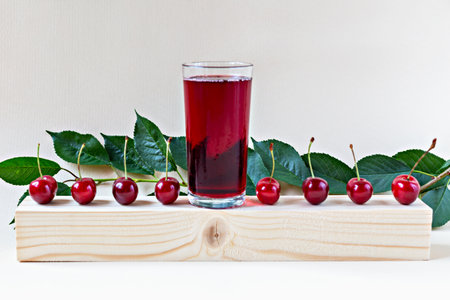 Glass of cherry juice with ripe cherries and green leaves on wooden block against light background. Ideal for ads, packaging, organic juice branding or healthy lifestyle concepts. Glass cherry juiceの写真素材