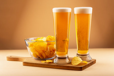 Two tall beer glasses with frothy heads and bowl of golden potato chips on wooden board against warm background. Concept of relaxation, delicious food and holiday. Two glasses beer with potato chipsの写真素材