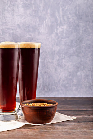 Two tall glasses of dark beer with foam served alongside roasted peanuts in a clay bowl. Rustic composition with wooden table and textured wall. Concept of authentic pub. Dark beer with rustic peanutsの写真素材